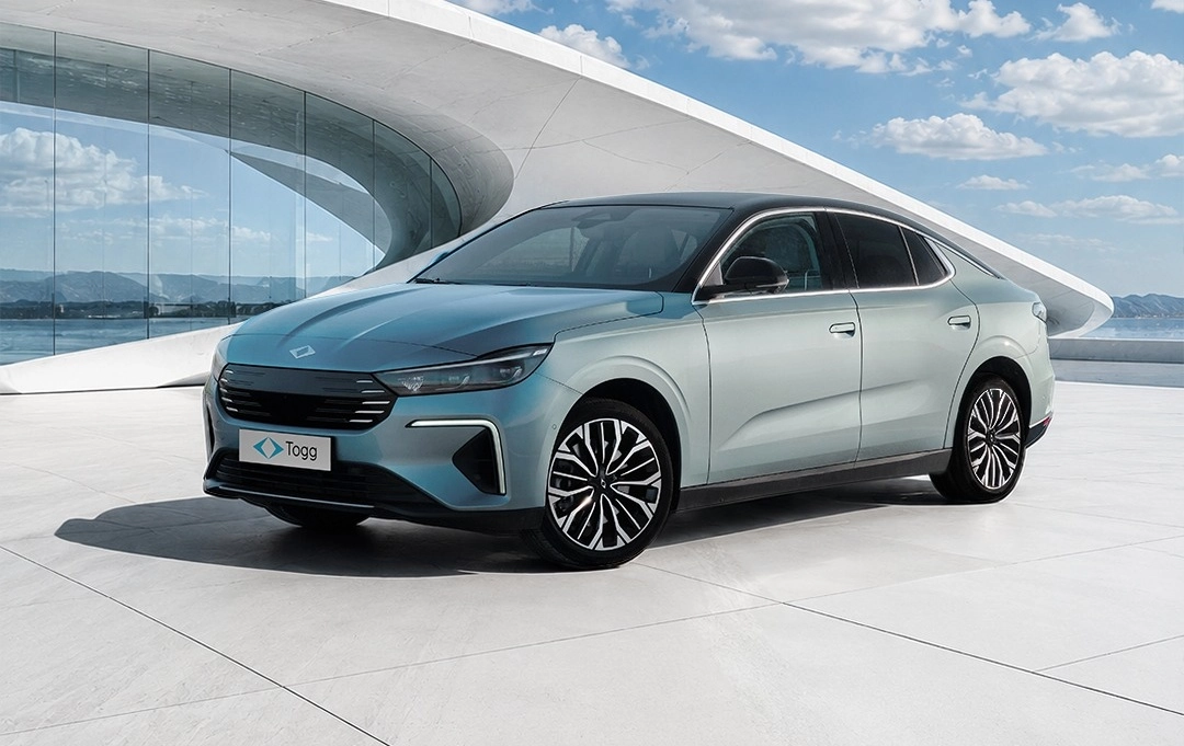 Mavi tonlarında, modern hatlara sahip Togg T10F elektrikli sedan otomobil, şık bir mimariye sahip camlı bir binanın önünde, geniş ve açık bir alanda parlak bir şekilde durarak 623 km menzil gibi önemli özelliklerini gözler önüne seriyor.