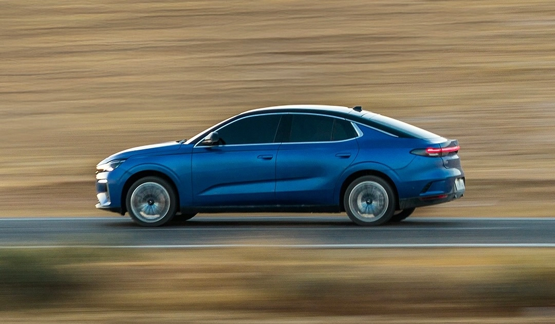 Mavi renkli, şık ve sportif hatlara sahip Togg T10F sedan elektrikli otomobil, hareket halinde yan profilden görüntülenmiş olup, bulanık arka plan ve dönen tekerlek detayları hız hissini pekiştiriyor.