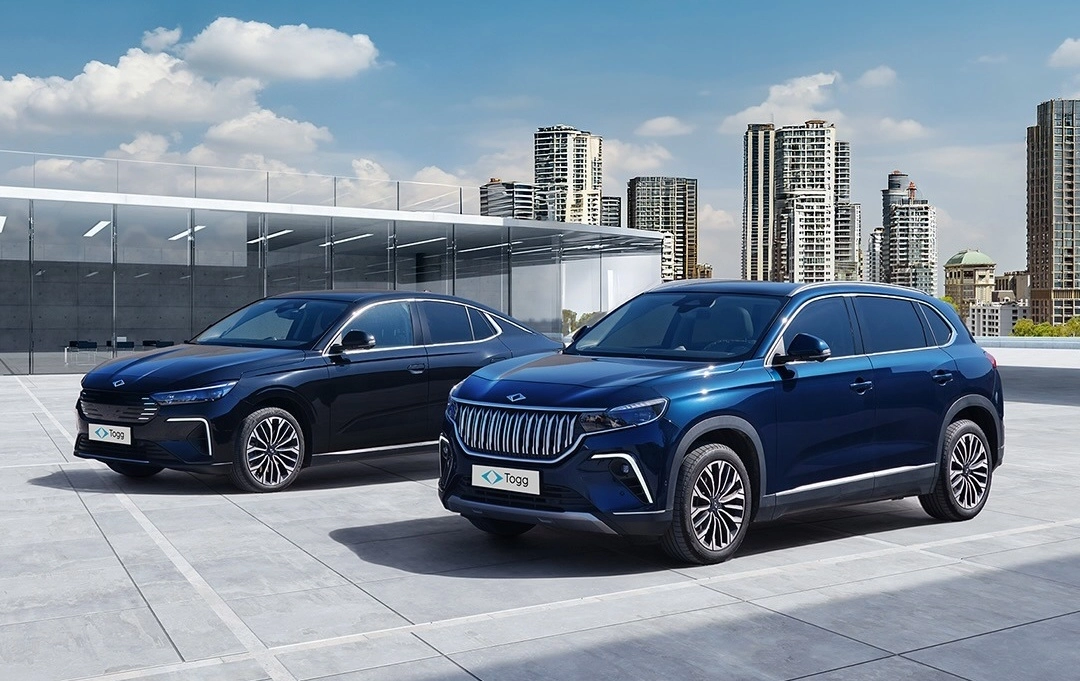 Güneşli bir günde, modern şehir binalarının ve cam cepheli bir yapının önünde şıkça duran, ön planda parlak mavi bir Togg T10X elektrikli SUV ile sol arkasında zarif siyah bir Togg T10F elektrikli sedan otomobil, Türk mühendisliğinin gücünü sergiliyor.