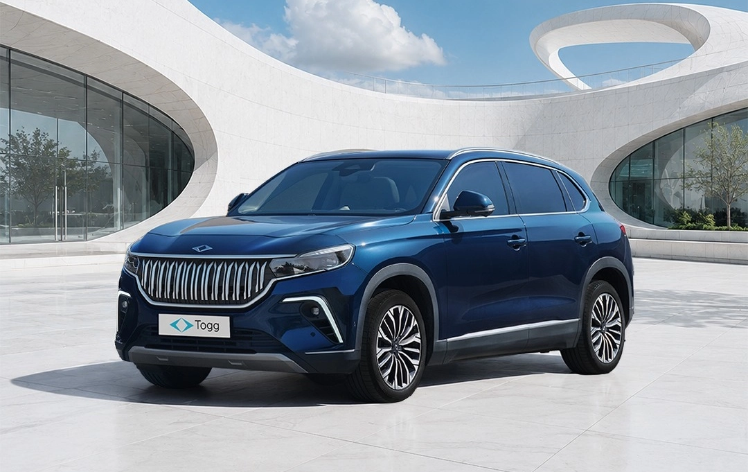 Koyu mavi renkli, ön tarafında Togg logosu bulunan şık Togg T10X elektrikli SUV otomobil, modern ve fütüristik kıvrımlı mimariye sahip beyaz bir binanın önündeki açık alanda park edilmiş, aydınlık bir günde duruyor.