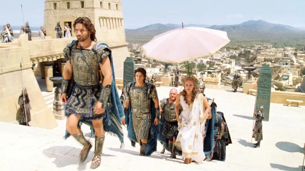 Truva (2004) filminden bir sahne: Antik zırhlar giymiş savaşçılar, bir merdivenden yukarı çıkarken aralarında beyaz bir elbise giymiş zarif bir kadın da yer alıyor. Kadının arkasında, güneşten korunması için büyük bir pembe şemsiye tutuluyor. Arka planda, antik bir şehir panoraması ve uzaklarda dağlar görünüyor. Sahne, Troya krallığını ve klasik Yunan atmosferini yansıtıyor.
