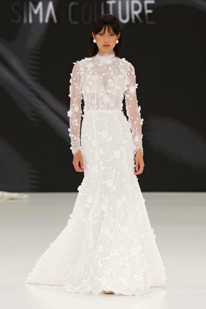 Üç boyutlu çiçek aplikelerle süslenmiş, transparan uzun kollu ve dik yakalı Sima Couture marka zarif ve romantik gelinlik modeli.
