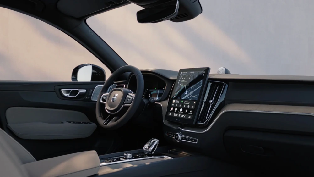 Volvo XC60 T8 AWD şarj edilebilir hibrit otomobilin kaliteli iç mekan tasarımını vurgulayan bu geniş açılı çekimde, yolcu koltuğundan görünen modern direksiyon simidi, üzerinde harita ve uygulama detayları bulunan büyük dikey multimedya ekranı ve şık vites kolu detayları öne çıkıyor.
