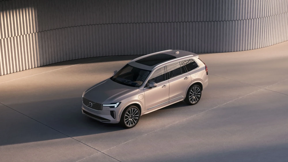 Açık renkli, modern ve zarif bir tasarıma sahip Volvo XC90 T8 AWD şarj edilebilir hibrit otomobil, modern bir yapının önünde beton zeminde park halinde dururken aracın dış tasarımını sergiliyor.
