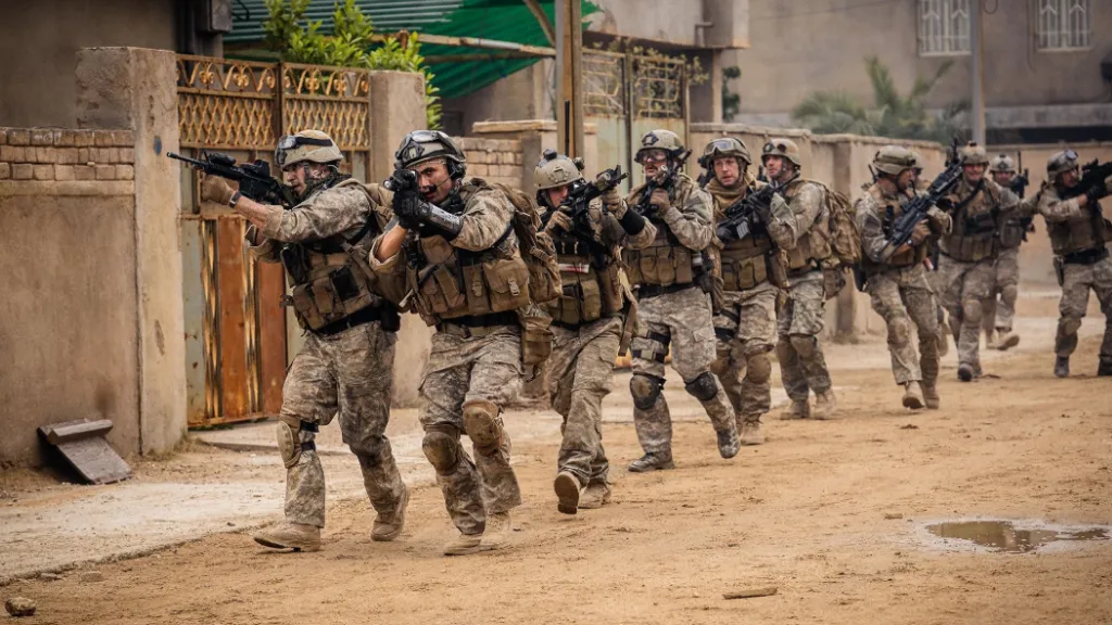 Warfare filminden modern savaş sahnesi: Tam teçhizatlı Amerikan askerleri, çöl benzeri bir şehirde sıralı ve dikkatli biçimde ilerliyor. Ellerinde tüfekler, üzerlerinde kamuflaj kıyafetler ve kasklar var. Arka planda Orta Doğu’yu andıran bir mahalle yer alıyor. Sahne taktiksel operasyon ve savaş ortamının ciddiyetini yansıtıyor.