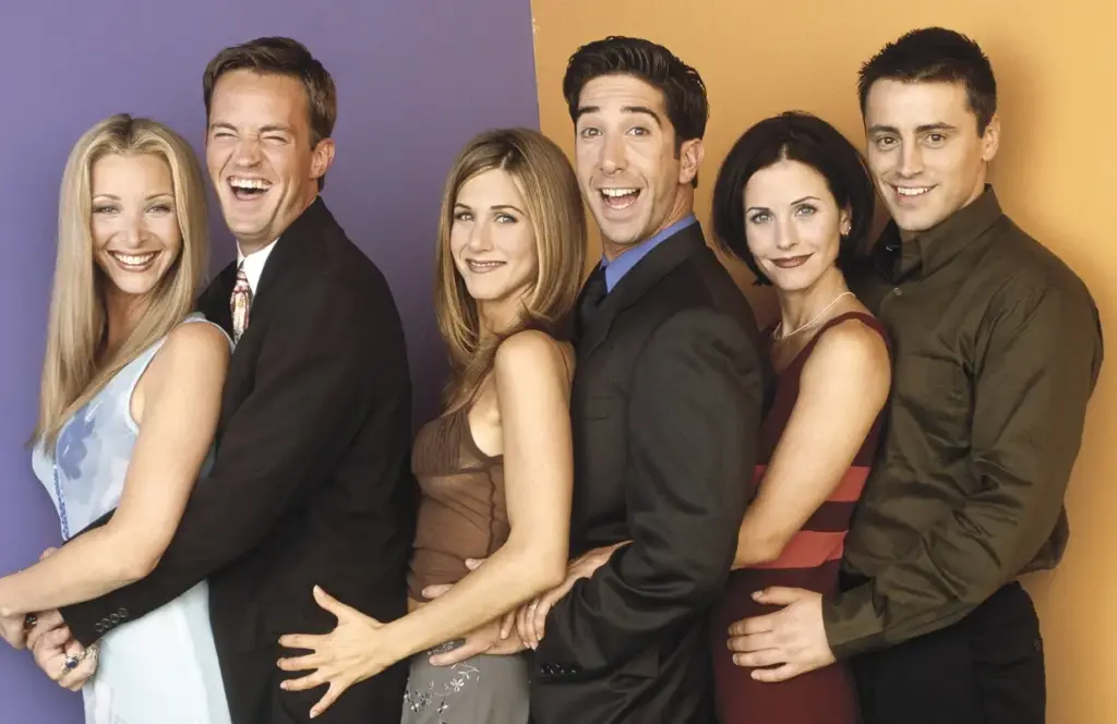Friends dizisinin ana karakterleri, arka arkaya sıralanmış şekilde poz verirken, 90’ların ikonik sitcom havasını yansıtan stüdyo çekimi.