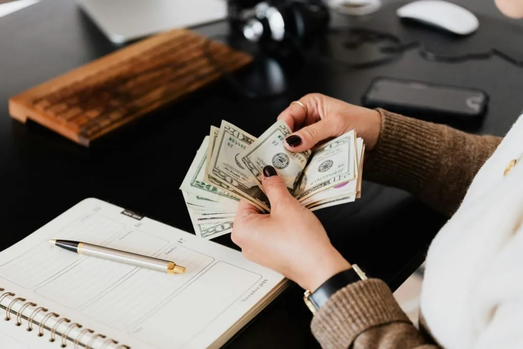 Bir kadın, elindeki dolar banknotlarını sayarken önünde bir ajanda ve kalem bulunuyor; yatırım planlaması yapılıyor izlenimi veriyor.