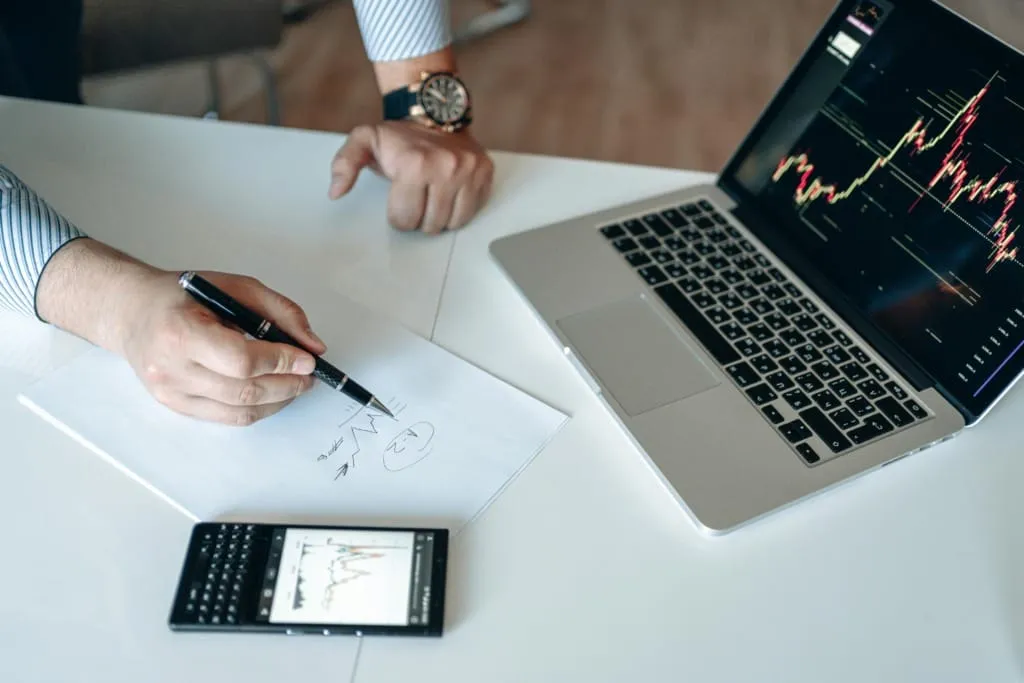 Bir erkek, masa başında kağıda finansal grafik çizerken, dizüstü bilgisayar ve hesap makinesinde yatırım verilerini analiz ediyor.