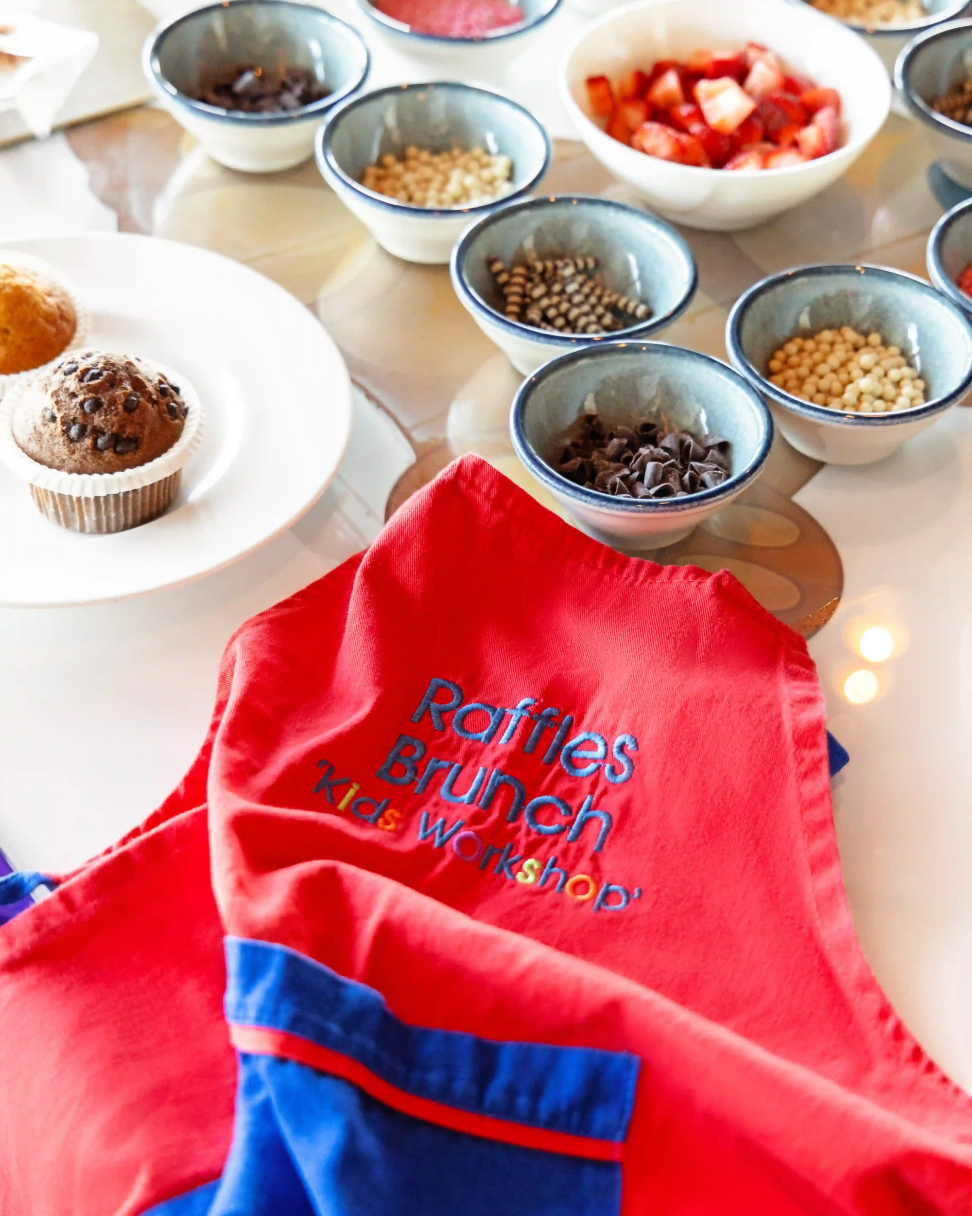 Beyaz bir tabakta duran iki çikolatalı muffin ile masada küçük kaselerde özenle sunulan doğranmış çilekler, çikolata parçacıkları ve çeşitli renkli şekerlemelerin yer aldığı, ön planda Raffles Brunch Çocuk Atölyesi yazısıyla bir aprona benzeyen kırmızı bir kumaşın görüldüğü bu görüntü, keyifli bir etkinliğin hazırlığını yansıtıyor.