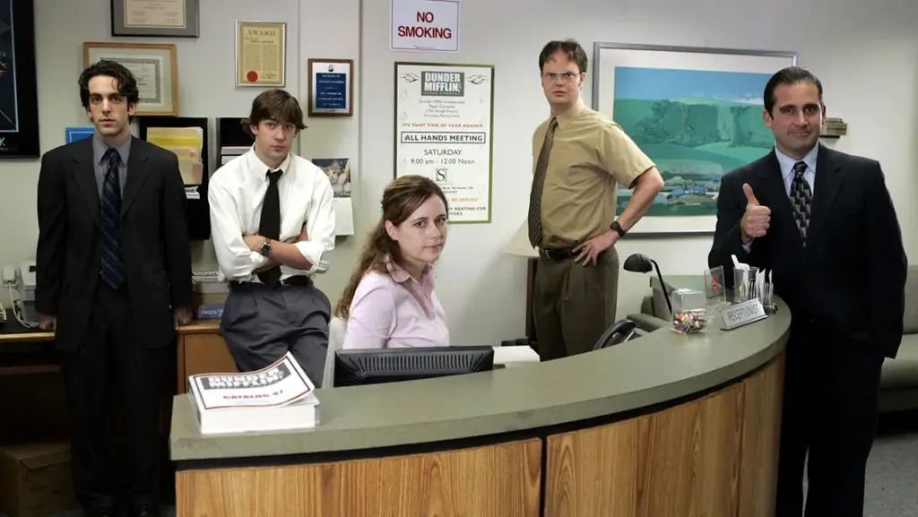 The Office dizisinin ana karakterleri Dunder Mifflin ofisinde resepsiyon masası çevresinde toplanmış, klasik işyeri atmosferi yansıtılıyor.