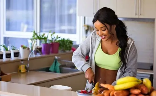 Mutfakta meyve doğrayarak sağlıklı öğün hazırlayan genç kadın; aktif yaşam, bitki bazlı beslenme ve dengeli rutin temalı modern wellness sahnesi.
