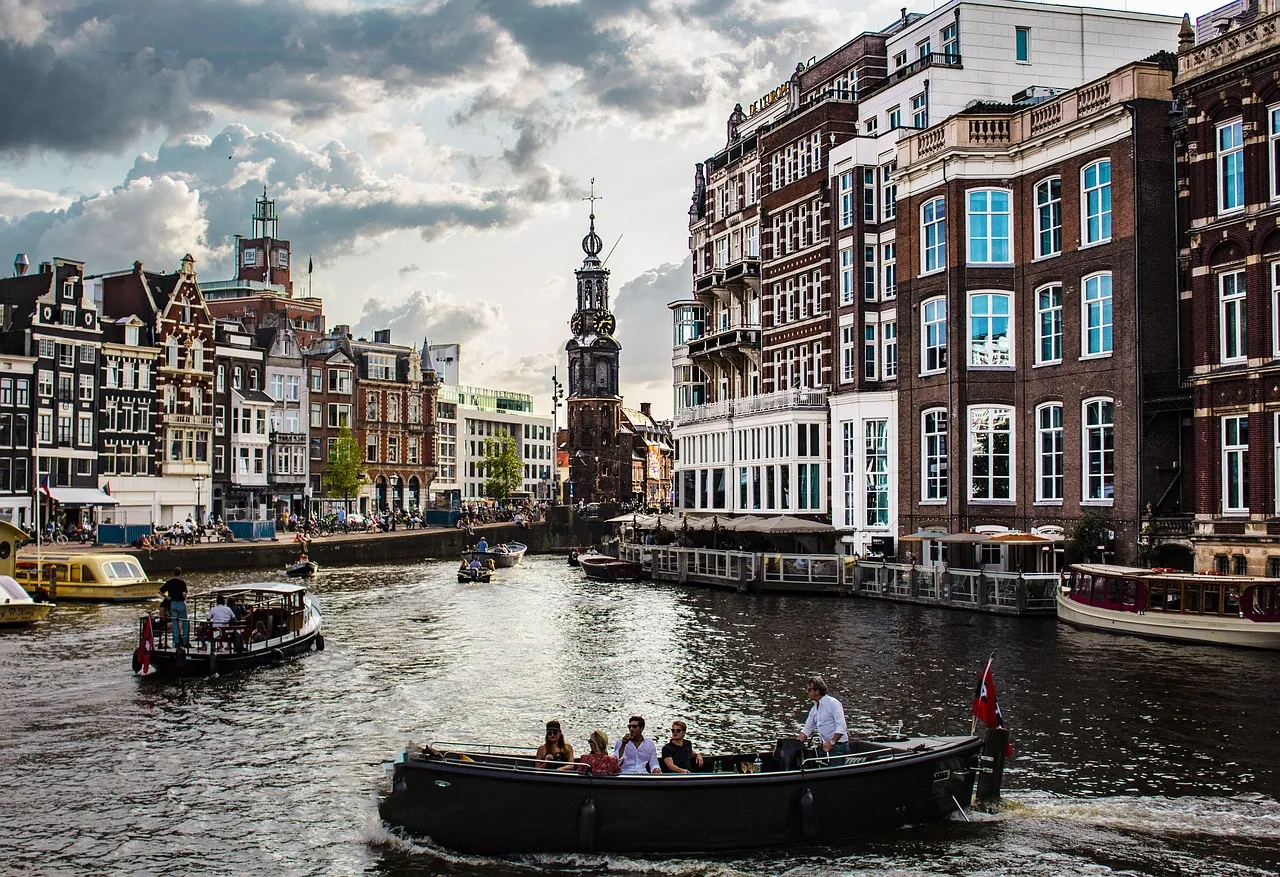Amsterdam’da tarihi kanal boyunca ilerleyen tekneler, arka planda dar cepheli tuğla binalar ve Westerkerk Kulesi, bulutlu gökyüzü altında şehir manzarası.