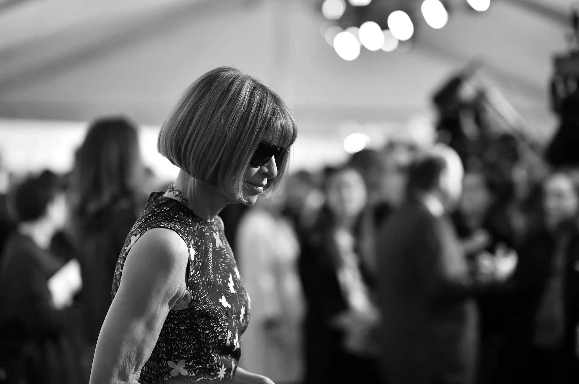 Siyah beyaz bir fotoğrafta, Vogue'un baş editörü ve Met Gala'nın önemli organizatörü Anna Wintour, karakteristik küt kesim saçları ve güneş gözlükleriyle, kolsuz, karmaşık desenli elbisesi içinde, New York'taki prestijli moda dünyası etkinliğinin kalabalık atmosferinde hafifçe yere doğru bakarken yandan profilden görünüyor.