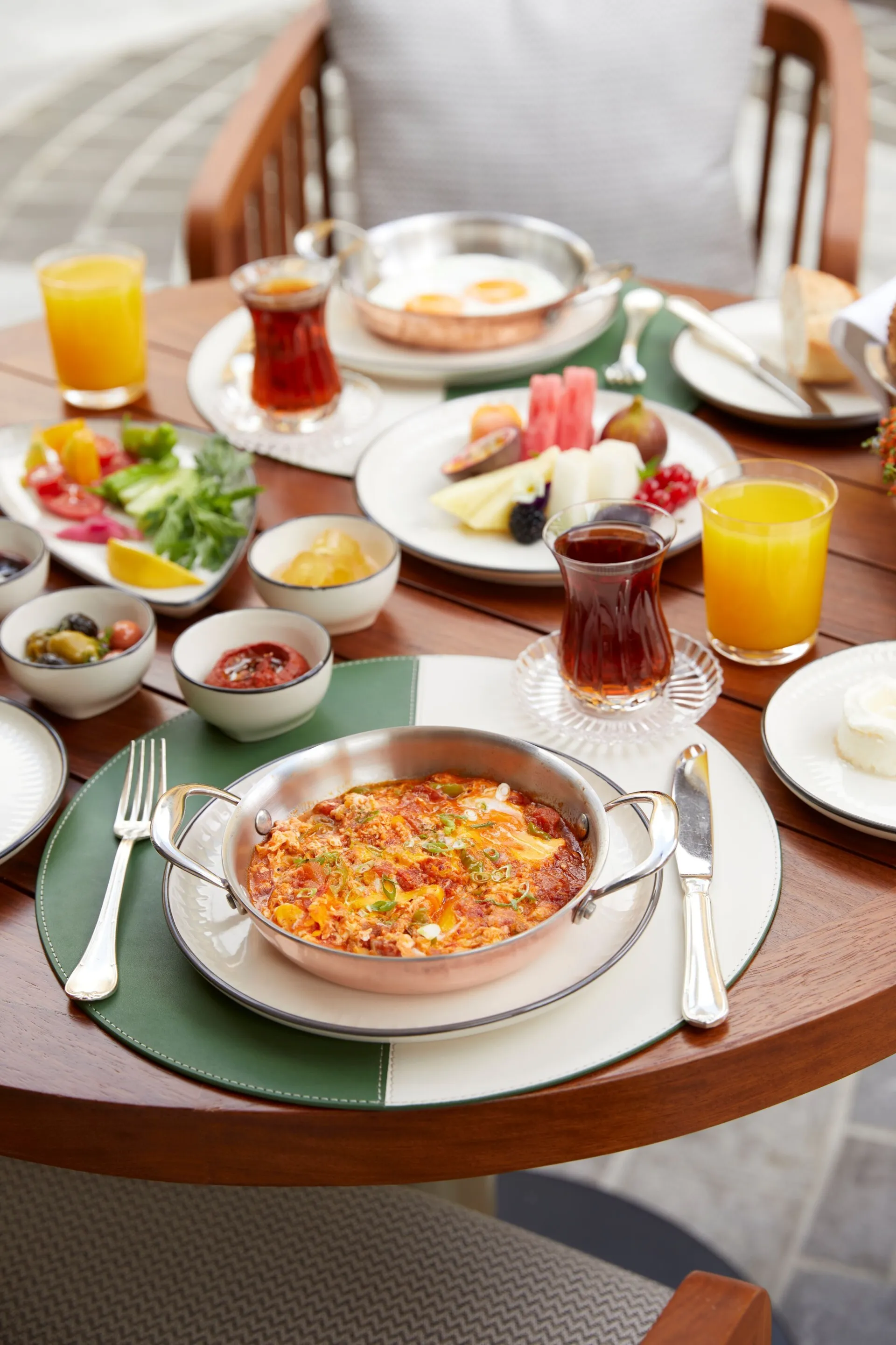 Anneler Günü brunch menüsünden menemen, sahanda yumurta, taze meyveler, zeytin, acuka ve salata gibi çeşitli lezzetlerin servis edildiği, portakal suyu ve çayların da eşlik ettiği şık bir ahşap masada özenle hazırlanmış zengin bir sofra görünümü.