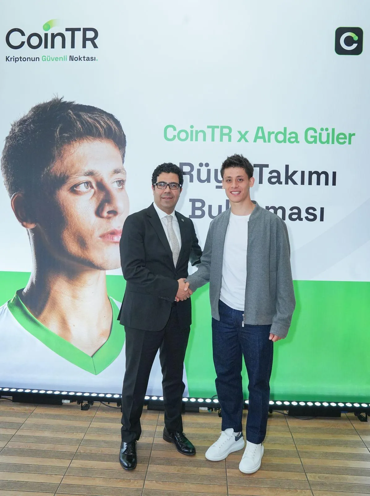 CoinTR x Arda Güler ve Rüya Takımı Buluşması yazılı, genç futbolcu Arda Güler'in büyük bir portre fotoğrafının da yer aldığı beyaz ve yeşil renkli bir pankart önünde, takım elbiseli bir yetkili ile Arda Güler gülümseyerek samimi bir şekilde el sıkışıyor.