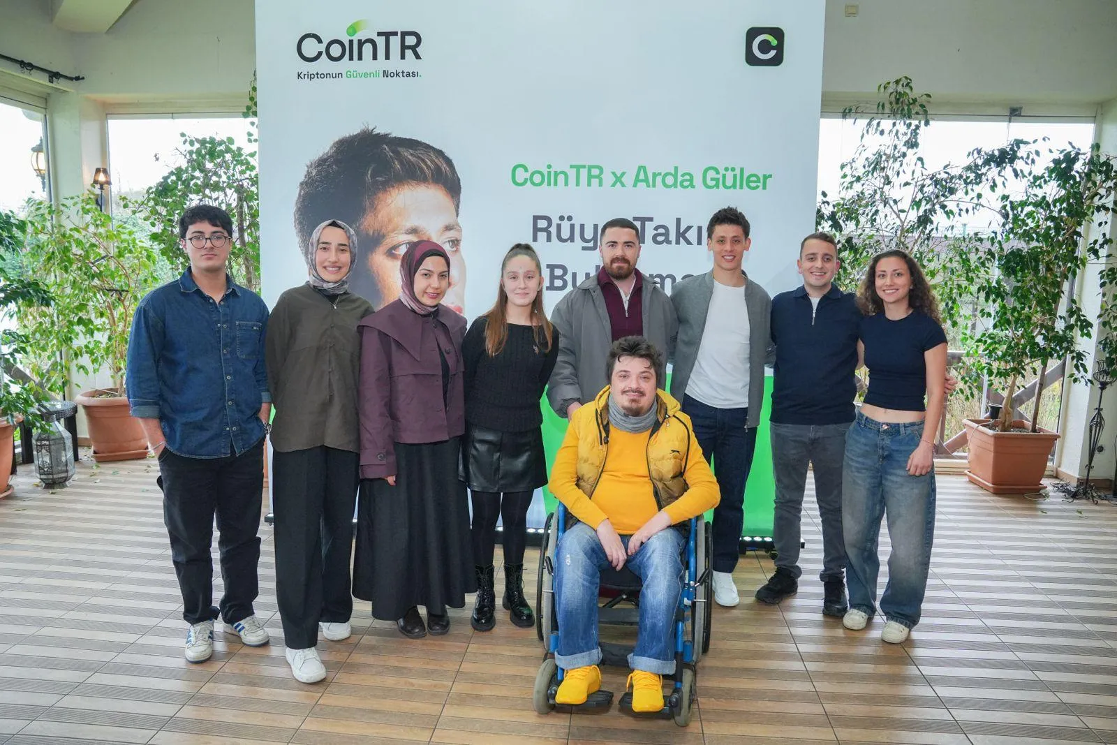 Sekiz ayakta duran ve bir tekerlekli sandalyede oturan toplam dokuz genç, arkalarında CoinTR logosu, Arda Güler'in başının hafifçe göründüğü büyük bir fotoğrafı ve Rüya Takımını Bulma etkinliği yazılarının yer aldığı beyaz pankartın önünde kameraya gülümseyerek poz veriyor.