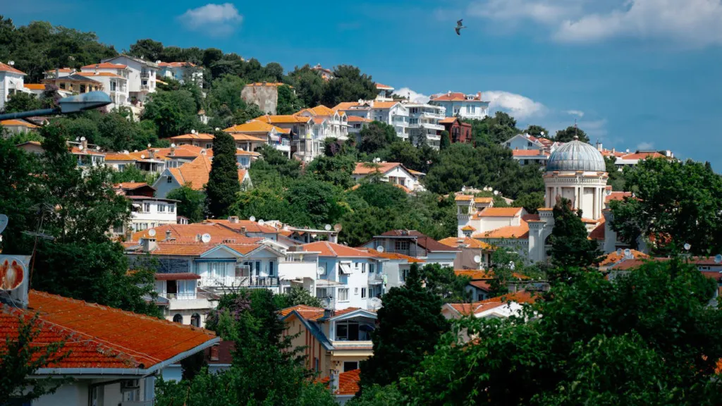 İstanbul'un Prens Adaları'ndan Burgazada'nın yemyeşil tepelerinde, kırmızı çatılı evlerin ve Aya Yani Kilisesi'nin kubbesinin berrak gökyüzünde süzülen bir martıyla huzurlu bir şekilde sıralandığı doğa kaçışı manzarası.