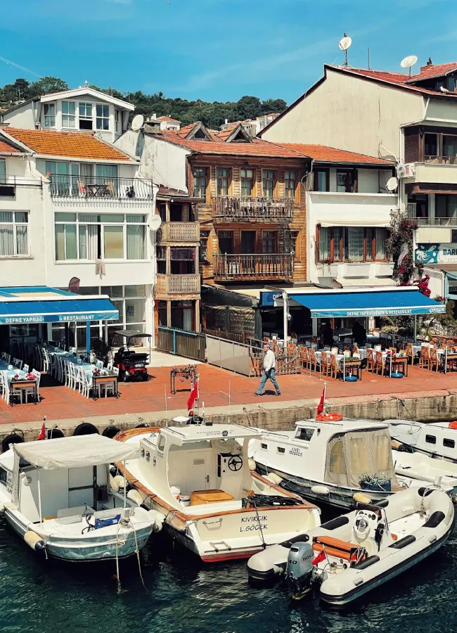 Burgazada sahilinde demirli teknelerin sıralandığı, mavi tenteli restoranların ve geleneksel evlerin bulunduğu dingin kıyı şeridi, yemyeşil tepelerin huzur veren manzarasıyla İstanbul'dan bir doğaya kaçış ve sakin bir rota sunuyor.