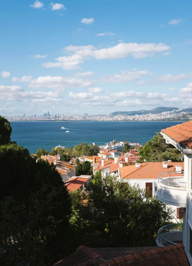 Büyükada'dan İstanbul siluetine doğru uzanan masmavi Marmara Denizi'nin üzerinden, kızıl kiremitli çatılar ve yemyeşil ağaçlarla kaplı adanın huzurlu doğasını gösteren bu görüntü, şehrin karmaşasından uzaklaşmak için ideal bir doğaya kaçış rotasını betimler.