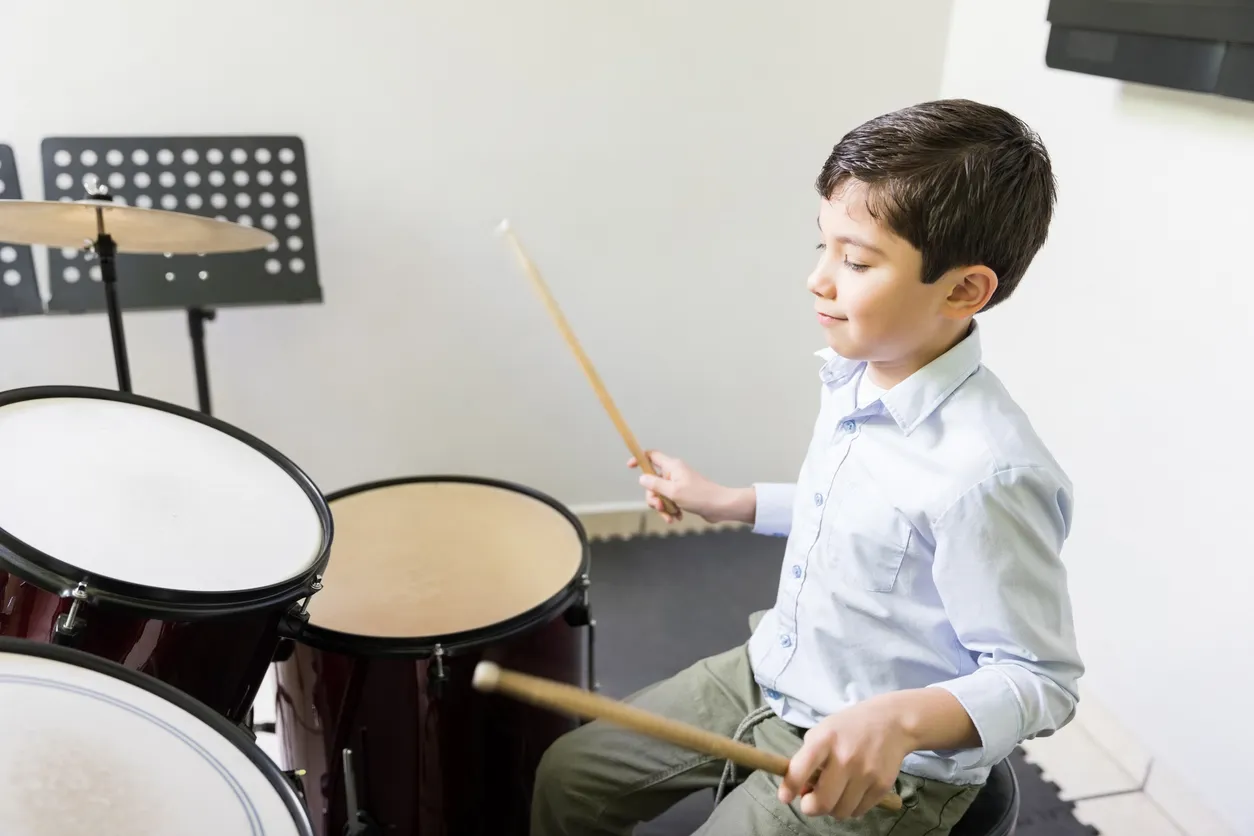 Müzik standının önünde oturmuş, mavi gömlekli bir çocuk elindeki bagetlerle bateri çalarken gözlerini kapatarak kendini ritmin akışına bırakmış, 23 Nisan çocuk etkinlikleri kapsamında sunulan bateri çocuk orkestrası gibi sanat dolu anların keyfini çıkarıyor.