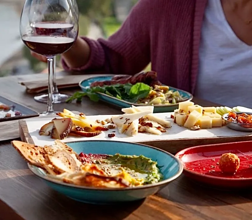 Bir Datça restoranında, gün batımı ışıkları altında zengin bir aperitif masası; dilimlenmiş ve küp şeklinde farklı peynir çeşitleri, cevizler, kurutulmuş meyveler, ekmek dilimleri, yeşil ve kırmızı tonlarda iki farklı dip sos, şarküteri ürünleri ve bir kadeh kırmızı şarapla dolu.