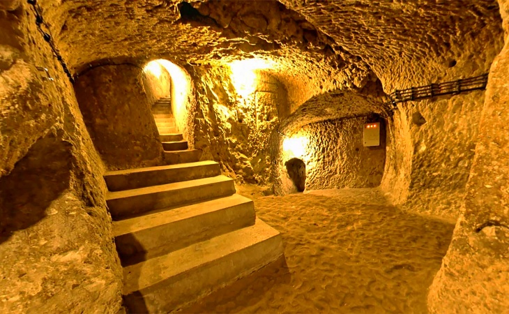 Kapadokya'daki Derinkuyu Yeraltı Şehri'nin loş sarı ışıklarla aydınlatılmış, kayadan oyma geniş bir geçidinde, sol tarafta yukarı doğru kıvrılan taş basamaklar ve sağda zeminde devam eden düz bir yol görülüyor, duvarlardaki pürüzlü doku ve modern tesisat izleri tarihi atmosferi modern dokunuşlarla harmanlıyor.