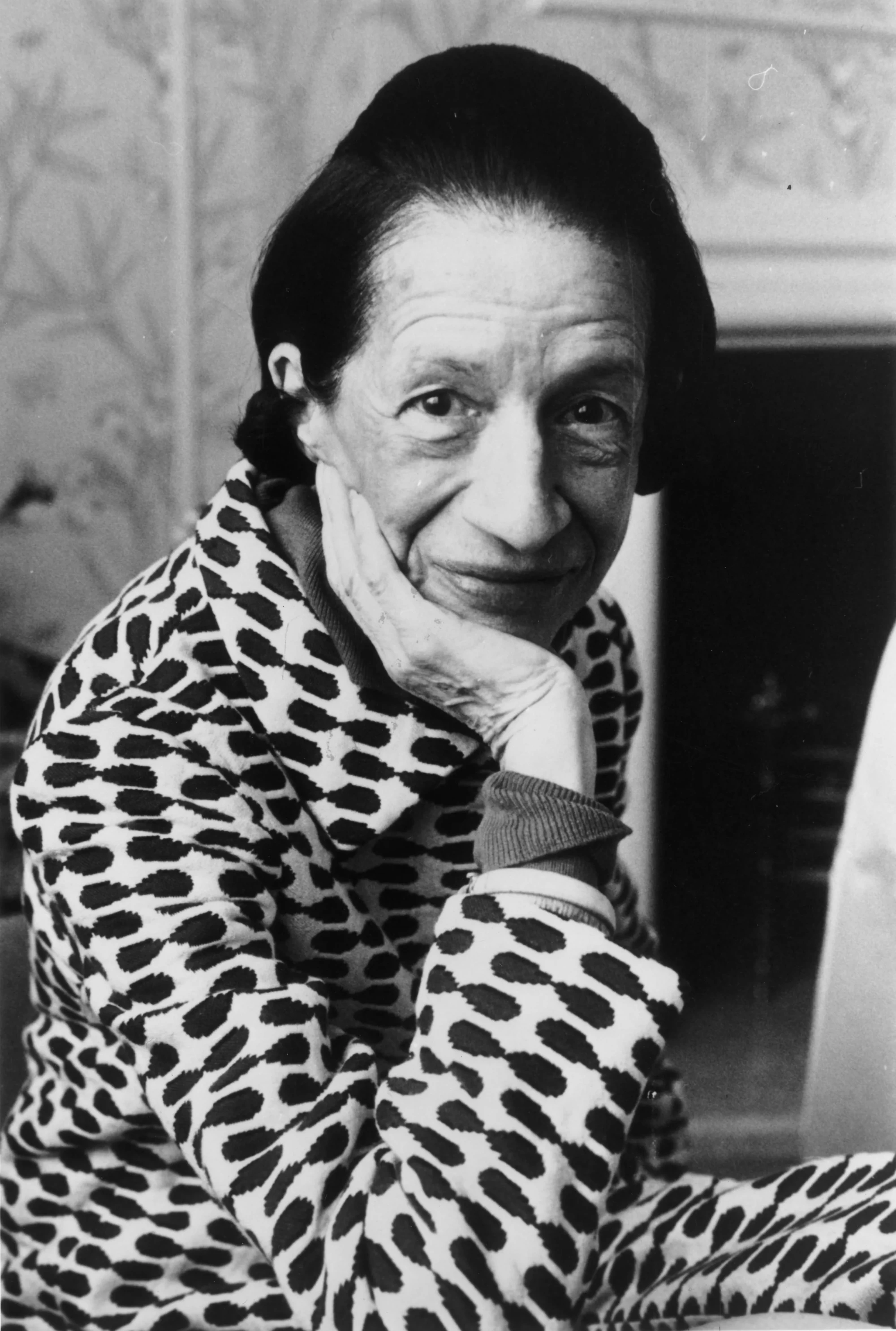 Met Gala'nın kültürel değişimine yön veren ünlü moda editörü Diana Vreeland, desenli bir ceketle, saçları geriye taranmış, hafifçe gülümseyerek çenesini eline dayamış bir şekilde, siyah beyaz portre fotoğrafında doğrudan izleyiciye bakıyor.