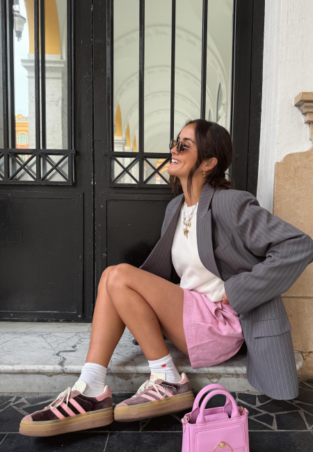 Şehirde bir binanın basamaklarında oturan şık bir kadın, çizgili gri blazer ceket, pembe şort ve gözlükleriyle kombinlediği kahverengi ve pembe renkli, retro esintili platform kadın sneaker ayakkabılarıyla neşeli bir şekilde gülümsüyor.