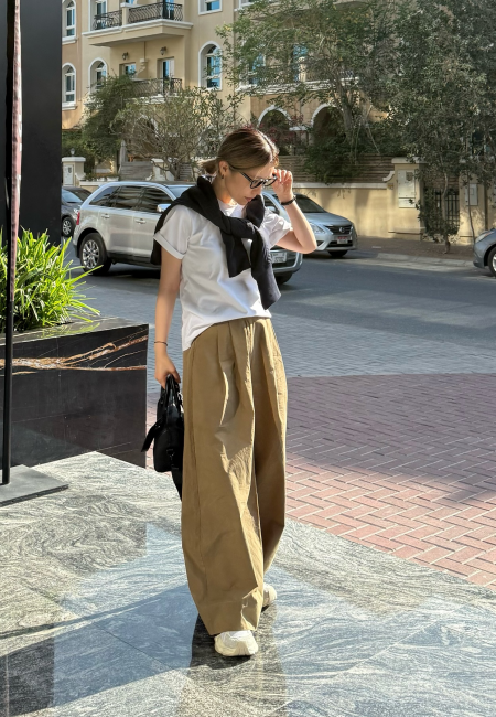 Güneşli bir havada, geniş paçalı haki pantolon ve beyaz tişört giyen, omuzunda siyah bir kazak olan genç bir kadın, taktığı güneş gözlüğünü ayarlarken duruşu ve ayağındaki kalın tabanlı açık renk trend kadın sneaker modelleri ile 2026'nın spor-şık stilini yansıtıyor.