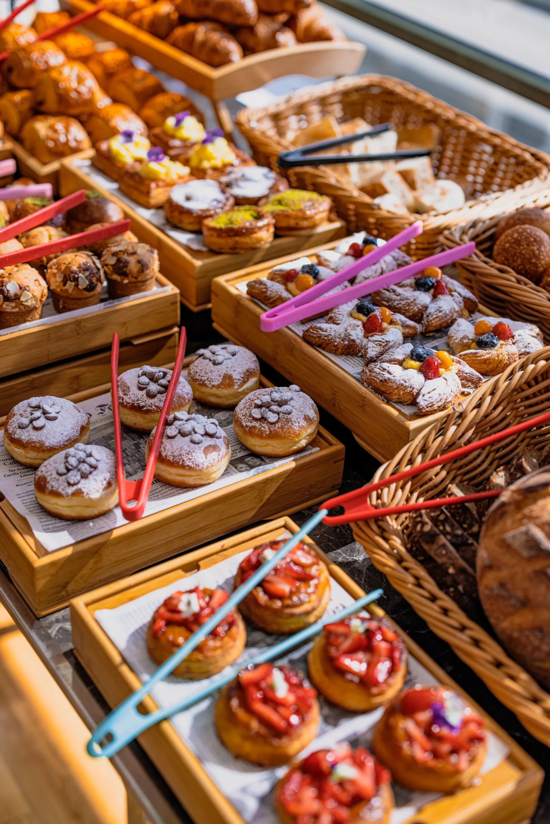 Parlak gün ışığı altında sergilenen bu zengin brunch büfesi, kruvasanlar, meyveli danimarka kekleri, bademli muffinler, pudra şekerli donutlar, taze çilekli tartlar ve hasır sepetlerde dilimlenmiş ekmekler gibi çok çeşitli tatlı ve tuzlu hamur işleriyle dolu olup, renkli servis maşalarıyla servis edilmeye hazır, davetkar bir kahvaltı ve öğle yemeği seçkisi sunuyor.