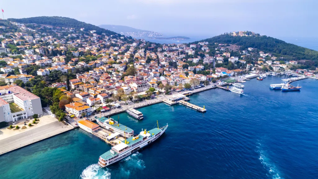 İstanbul’da doğaya kaçış için ideal bir huzurlu rota olan Heybeliada'nın kuşbakışı manzarasında, yemyeşil tepelerin eteklerine yayılan kırmızı çatılı evler, masmavi denizde hareketli iskeledeki vapurlar ve turkuaz sular dikkat çekiyor.