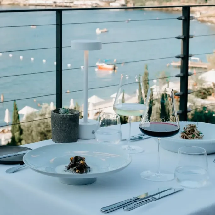 Bodrum'un seçkin restoranlarından birinde, masmavi Ege Denizi manzarasına nazır beyaz örtülü zarif bir masada, özenle hazırlanmış modern yemekler, kırmızı ve beyaz şarap kadehleri ile şık bir masa lambasının eşlik ettiği, detaylara önem veren bir gastronomi deneyimi.