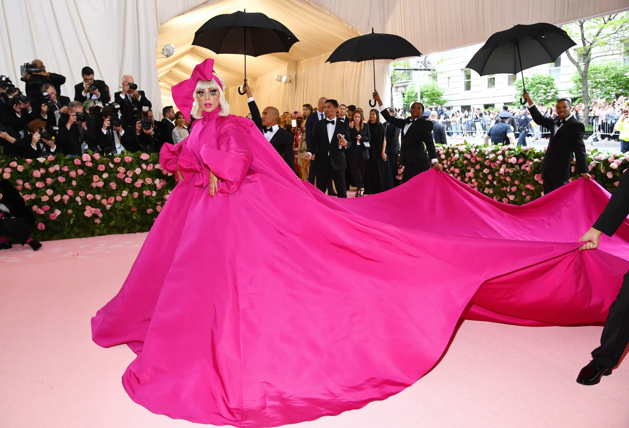 Lady Gaga, moda dünyasının en prestijli etkinliklerinden biri olan Met Gala'da, dramatik makyajı ve başındaki büyük fiyonkla dikkat çeken, uzun kuyruğu iki kişi tarafından taşınan ikonik fuşya rengi kabarık elbisesiyle etrafı fotoğrafçılarla çevrili pembe halıda yürüyor.