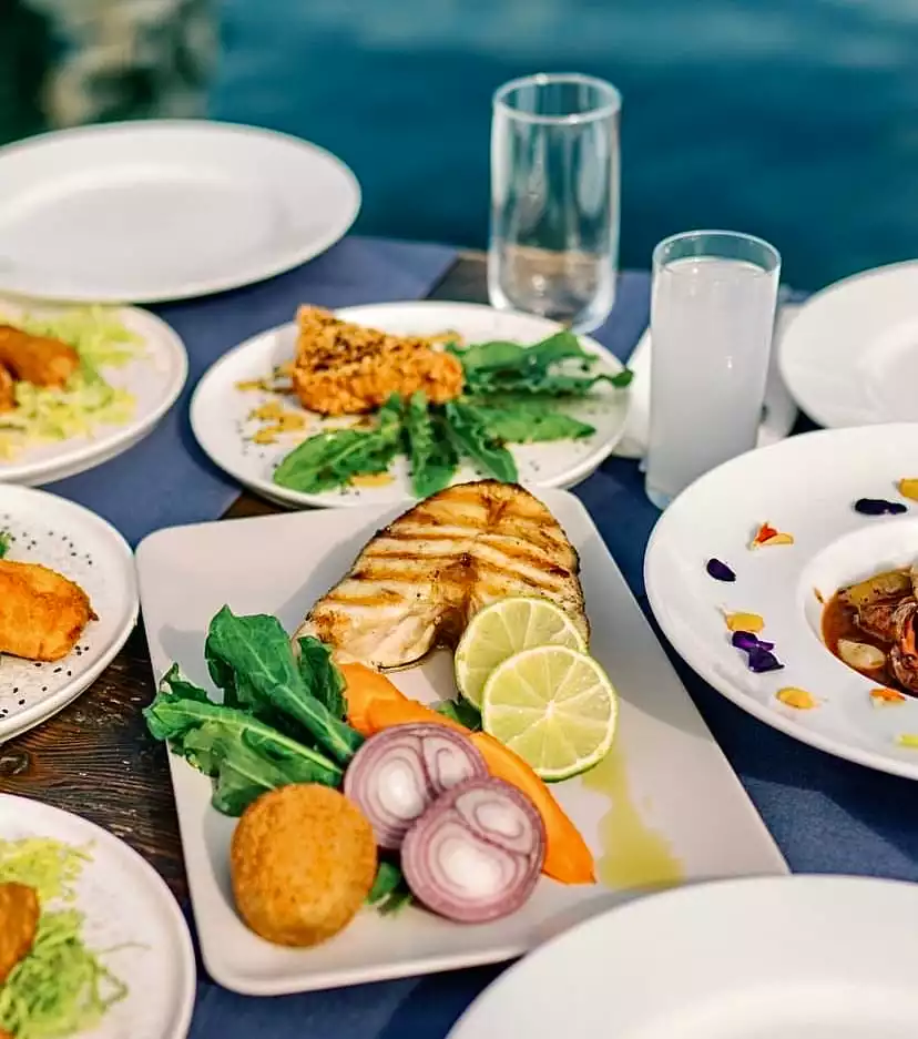 Mavi yolculuk keyfiyle uyumlu olarak, ızgara balık, taze roka, kırmızı soğan, havuç ve misket limon dilimleriyle bezenmiş ana deniz ürünleri tabağı; yanında mezeler, rakı bardağı ve huzur veren mavi deniz manzarasıyla zengin bir sofrayı gözler önüne seriyor.