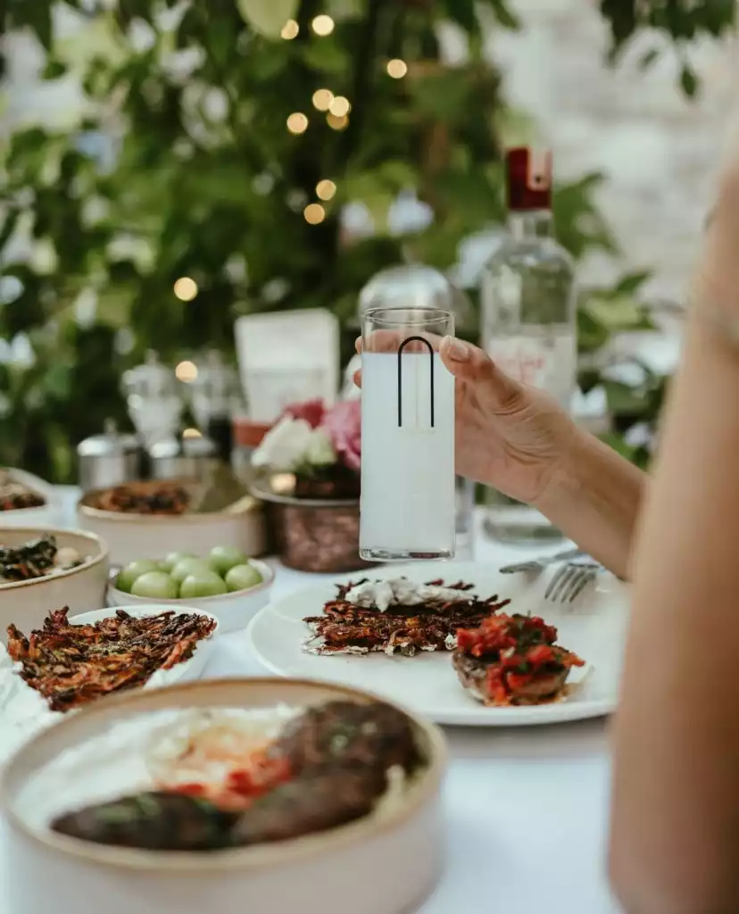 Bir elde tutulan, üzerinde minimalist siyah bir desen olan buzlu içecek dolu uzun bir bardak, arkaplanda ışıklı yeşil bitkilerle çevrili şık bir masada sunulan zeytinyağlılar, mücverler ve köfteler gibi geleneksel lezzetlerle dolu çeşitli meze tabaklarının ortasında keyifli bir Mavi Yolculuk yemeği anını yansıtır.
