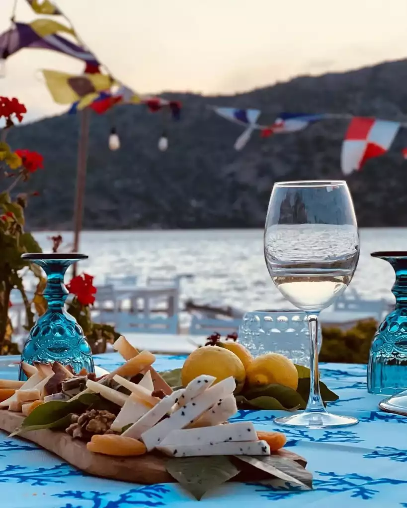 Denize nazır, mavi desenli örtü serili bir masada, peynir çeşitleri, ceviz, kuru kayısı ve limonlarla dolu ahşap bir sunum tabağı ile şık kadehler, mavi yolculuk temalı bir sofrayı oluşturuyor; arka planda ise renkli deniz bayrakları ve dağlık kıyı şeridinin huzurlu manzarası eşliğinde günbatımı ışıkları yansıyor.
