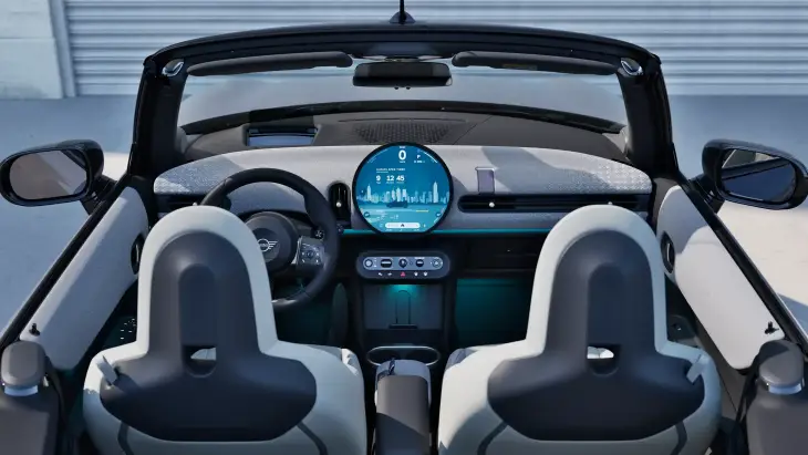 Yeni MINI Cooper Cabrio'nun üstü açık iç mekânında, şehir manzaralı dairesel OLED ekran ve minimalist kokpit tasarımıyla dijital konfor ve özgürlük hissi birleşirken, şık koltuklar ve mavi ambiyans aydınlatması modern bir sürüş deneyimi sunuyor.