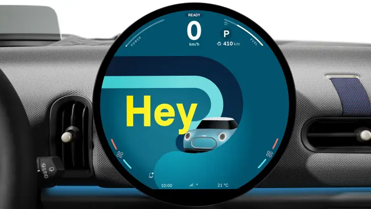 Yeni MINI Cooper Cabrio'nun iç mekanında, ortada yer alan ve üzerinde büyük sarı Hey yazısı, küçük bir araç ikonu, hız göstergesi ile 410 km menzil bilgisini gösteren dairesel OLED ekran, aracın dijital konforunu ve sesli asistan özelliğini vurgularken, dokulu gri gösterge paneli ve havalandırma menfezleri modern iç tasarımı tamamlıyor.