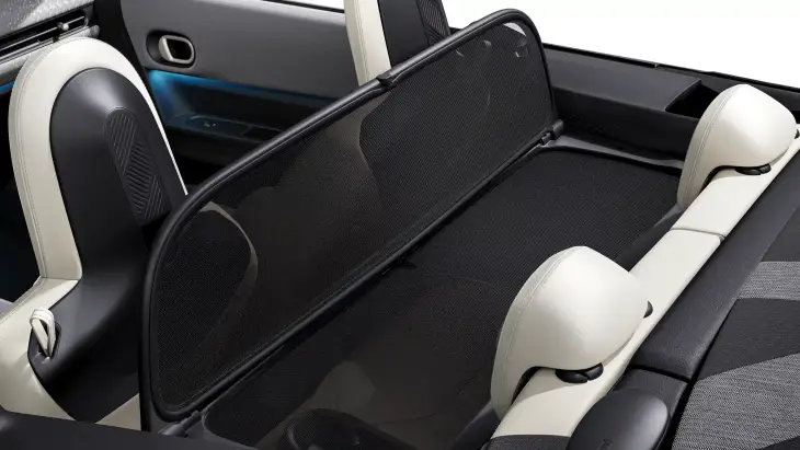 Yeni MINI Cooper Cabrio'nun bej detaylı iç mekanında, açık havada sürüş deneyimini daha konforlu kılan ve rüzgarı kesen siyah fileli rüzgar koruyucu, ön ve arka koltuklar arasına gerilmiş olarak görülüyor, bu da her an özgür bir sürüş vadettiğini gösteriyor.