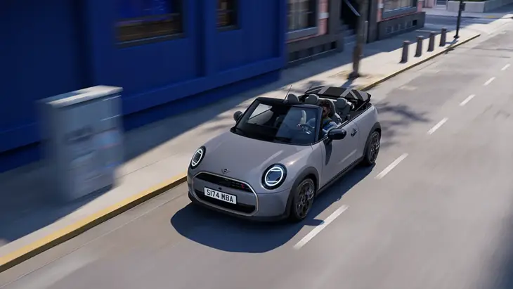 Şehir sokaklarında dinamik bir şekilde ilerleyen, tavanı açık gri renkli Yeni MINI Cooper Cabrio, parlak LED farları ve şık hatlarıyla dikkat çekerken, içindeki yolcularla özgür bir sürüş deneyimi sunan hareketli bir sahneyi betimliyor.
