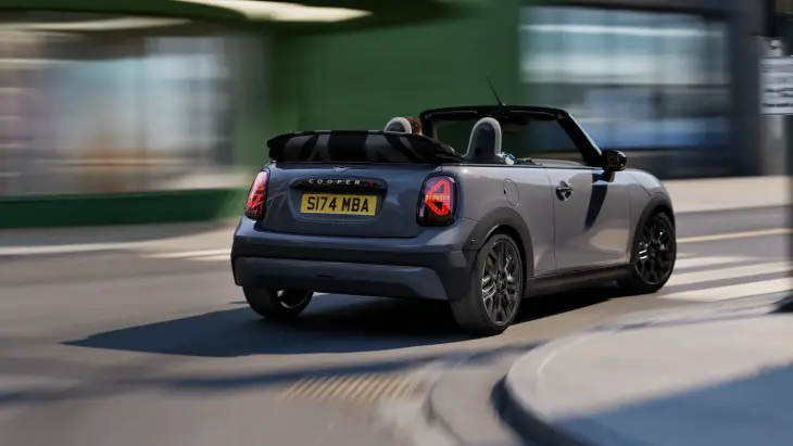 Şehirde sağa viraj alırken arkadan görünen, üstü açık gri Yeni MINI Cooper Cabrio, ikonik tasarımı ve yolcularıyla rüzgarın tadını çıkararak özgür bir sürüşün keyfini yaşatıyor gibi yolda hızla ilerliyor.