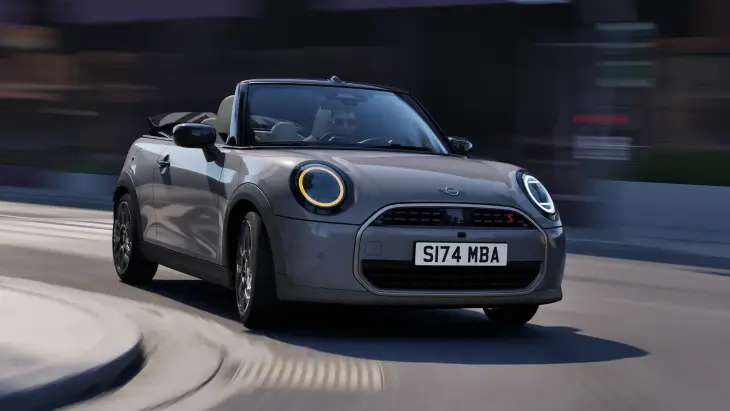 Yeni MINI Cooper Cabrio'nun ikonik tasarımını ve şehir sokaklarında sunduğu özgür sürüş deneyimini vurgulayan, gri renkli üstü açık bir MINI Cooper S modelinin virajlı yolda hızla ilerlerken çekilmiş dinamik ön-yan görüntüsü, aracın çevikliğini ve modern estetiğini başarılı bir şekilde yansıtıyor.
