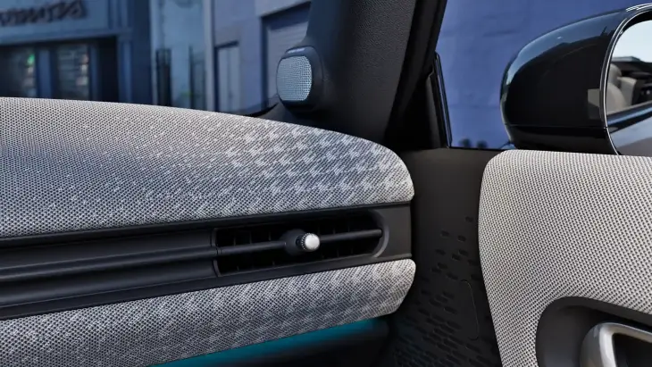 Yeni MINI Cooper Cabrio'nun iç mekanında, şık dokulu açık gri kumaş kaplı ön konsol ve kapı panelinin detaylı görünümü; minimalist tasarımlı havalandırma ızgarası, ambiyans aydınlatması ve hoparlör ile modern bir özgür sürüş deneyimini yansıtıyor.