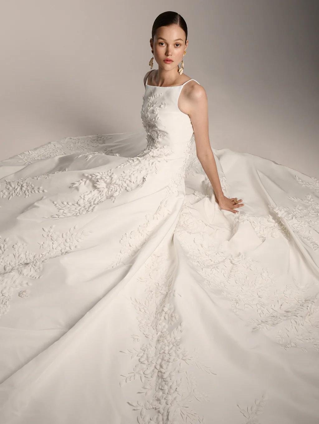Oscar de la Renta 2026 gelinlik modelleri koleksiyonundan, zarif ve heykelsi duruşuyla dikkat çeken, ince askılı kare yaka beyaz bir gelinlik giyen model yerde oturuyor; elbisenin volümlü etekleri ve bedeni, baştan sona uzanan zarif üç boyutlu çiçek ve yaprak aplikeleriyle büyüleyici bir şekilde bezeli.