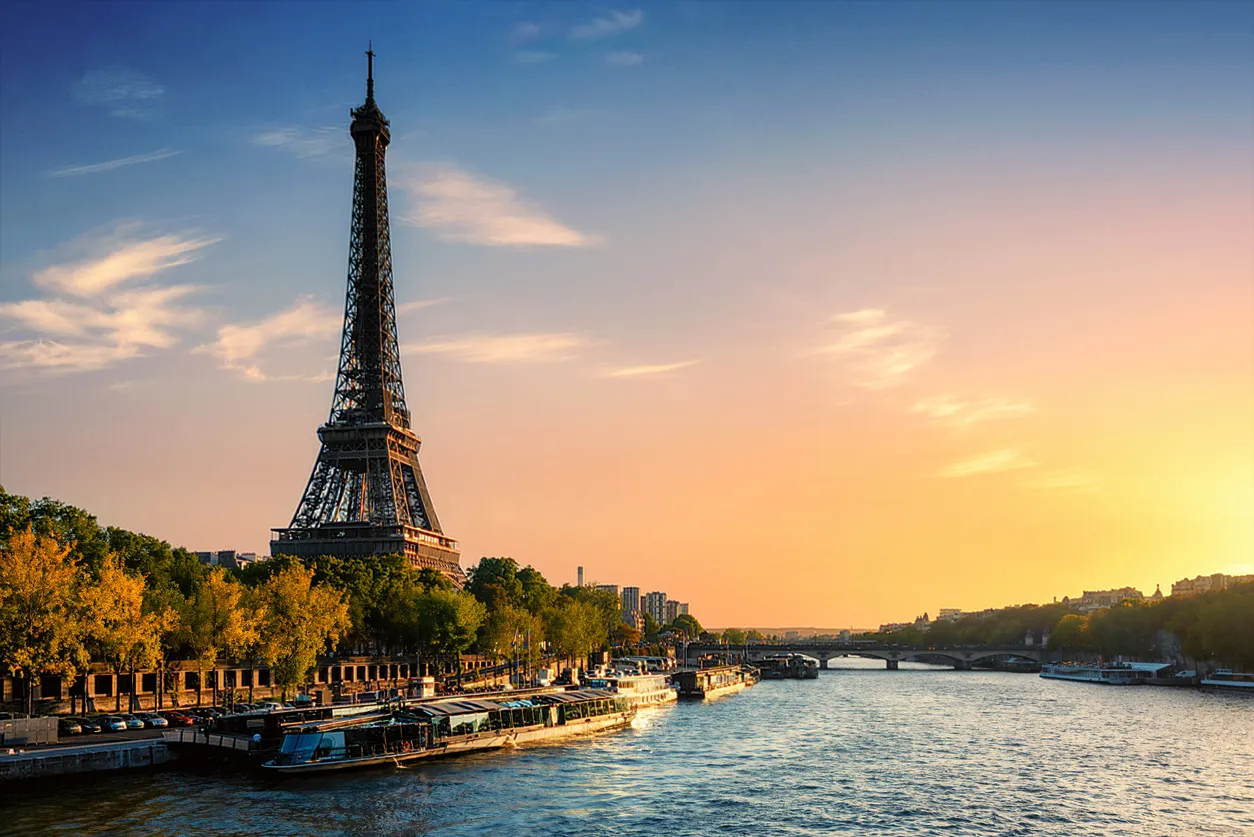 Eyfel Kulesi, gün batımının altın rengi ışıkları altında, Paris'in ikonik Sen Nehri'nde sıralanmış turist tekneleri ve yemyeşil ağaçlarla çevrili büyüleyici şehir siluetini ortaya koyan bir görüntü.