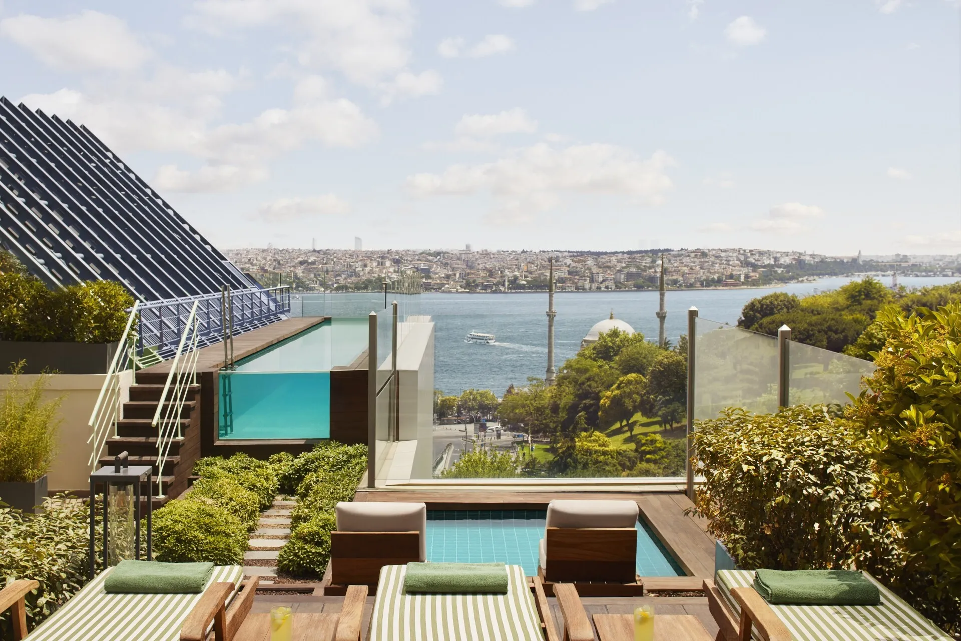 Ritz-Carlton İstanbul'un Boğaz'a nazır lüks terasında, çizgili şezlonglar ve cam kenarlı havuzun gerisinde, şehir panoraması, tarihi bir cami ve sol yanda modern mimari bir öğe ile Bosphorus'un nefes kesici manzarası uzanıyor.