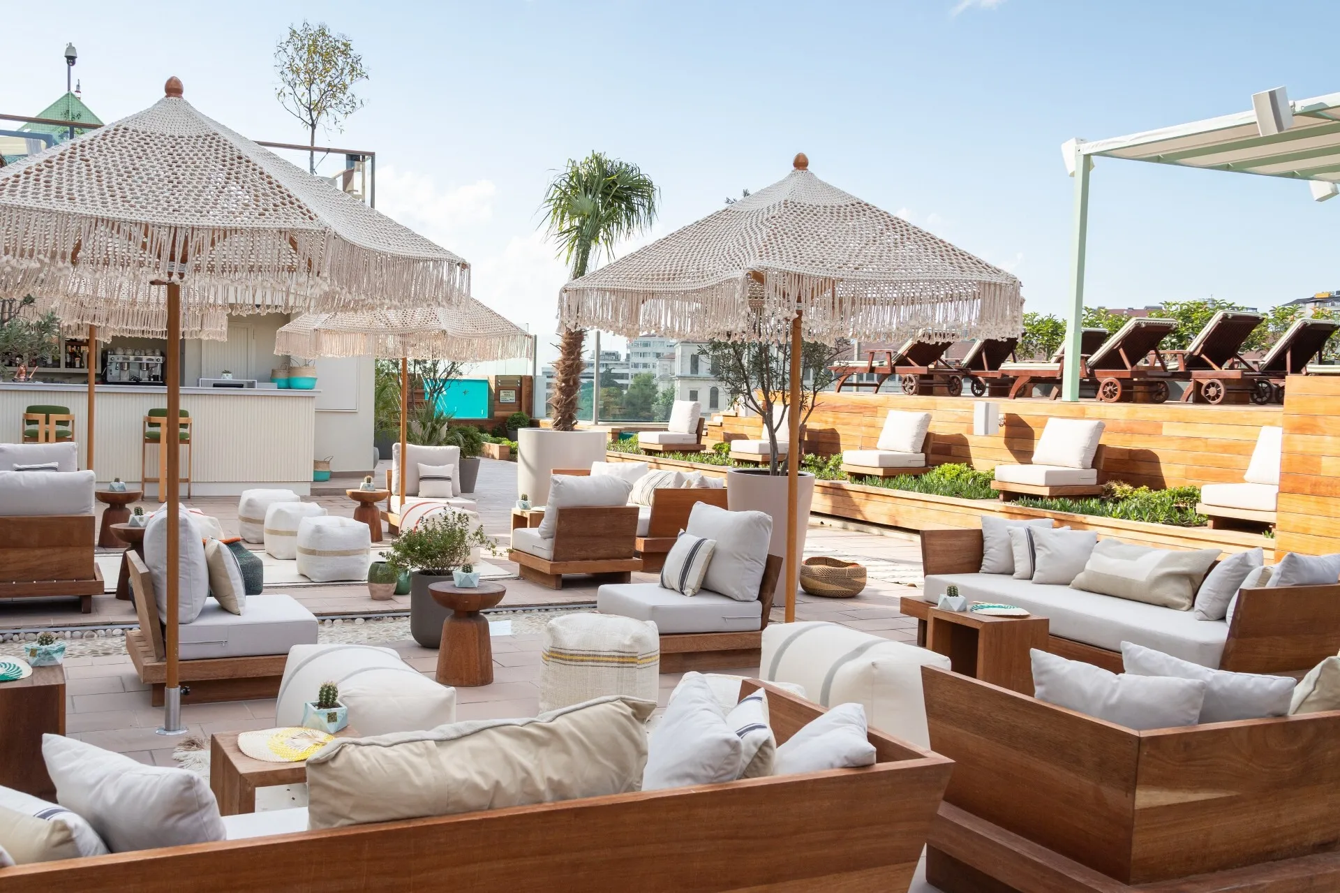 Ritz-Carlton otelinin geniş çatı terasında, bej ve beyaz tonlarda minderli ahşap koltuklar, puflar, dekoratif makrome şemsiyeler, arka planda bir bar ve güneşlenme alanındaki şezlonglar ile zarif ve davetkar bir açık hava ortamı göz alıcı bir şekilde düzenlenmiştir.