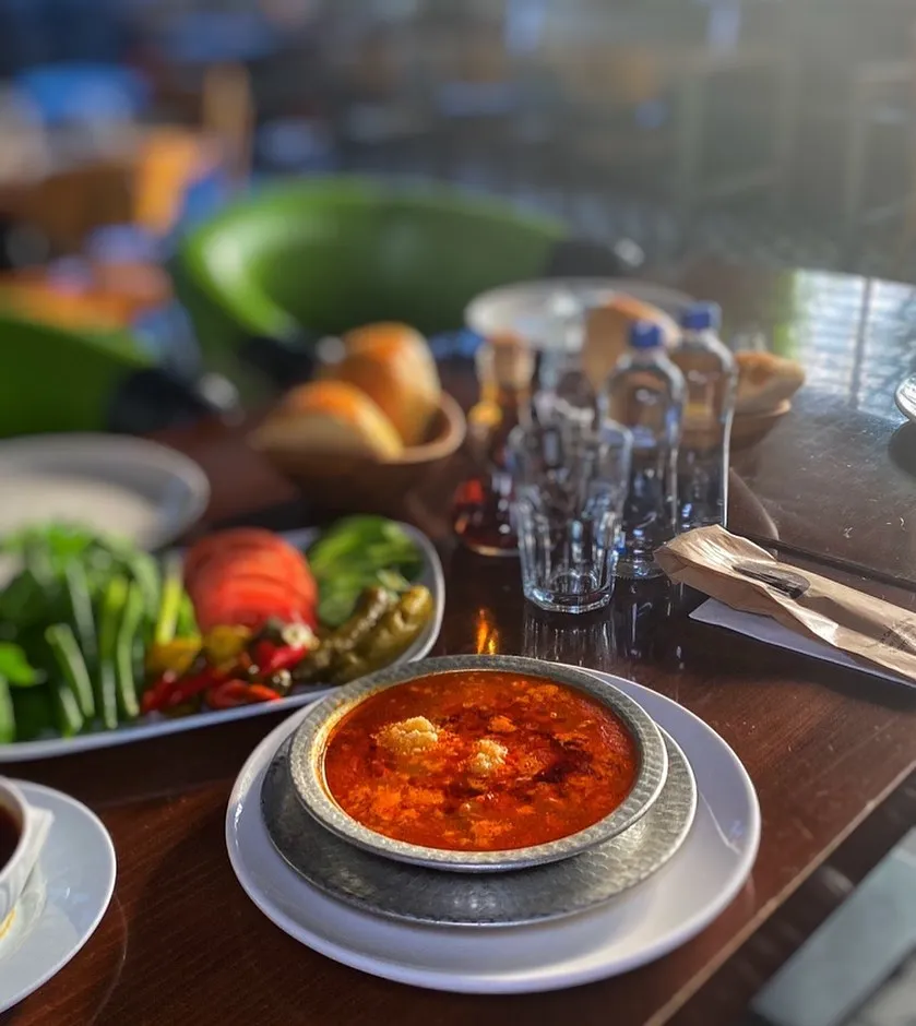 Etiler Sarıhan Gusto’da servis edilen kırmızı renkli kelle paça çorbası, yanında yeşillikler ve turşularla birlikte.