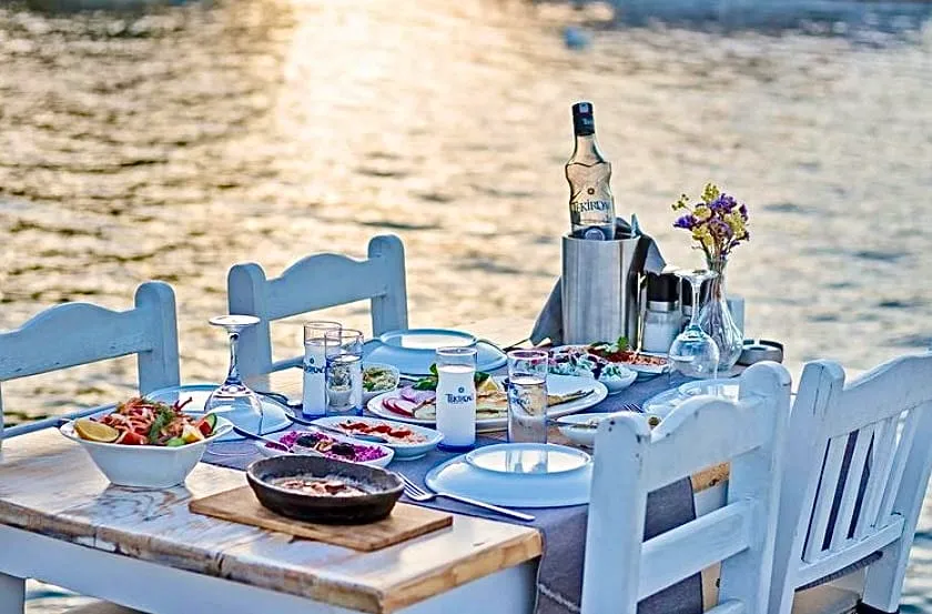 Gün batımı ışıklarında parlayan denize nazır bir Selimiye-Bozburun restoranında, üzerinde çeşitli Türk mezeleri, taze salata, bir şişe rakı ve süslü çiçeklerin bulunduğu, beyaz ahşap sandalyelerle çevrili, keyifli bir akşam yemeği için hazırlanmış ahşap masa.