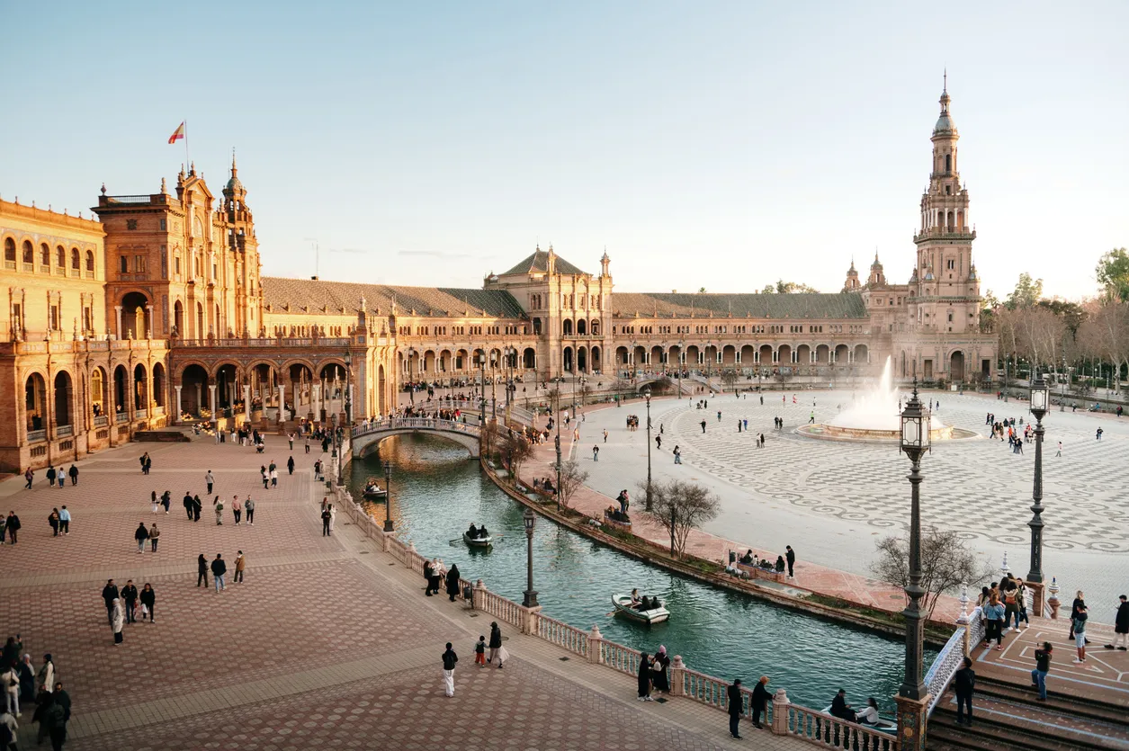 İspanya Sevilla’daki Plaza de España meydanı; yarım daire formundaki tarihi yapı, köprüyle geçen kanal, küçük sandallar ve ortadaki büyük fıskiye çevresinde dolaşan ziyaretçiler, gün batımı ışığında geniş açı şehir manzarası.